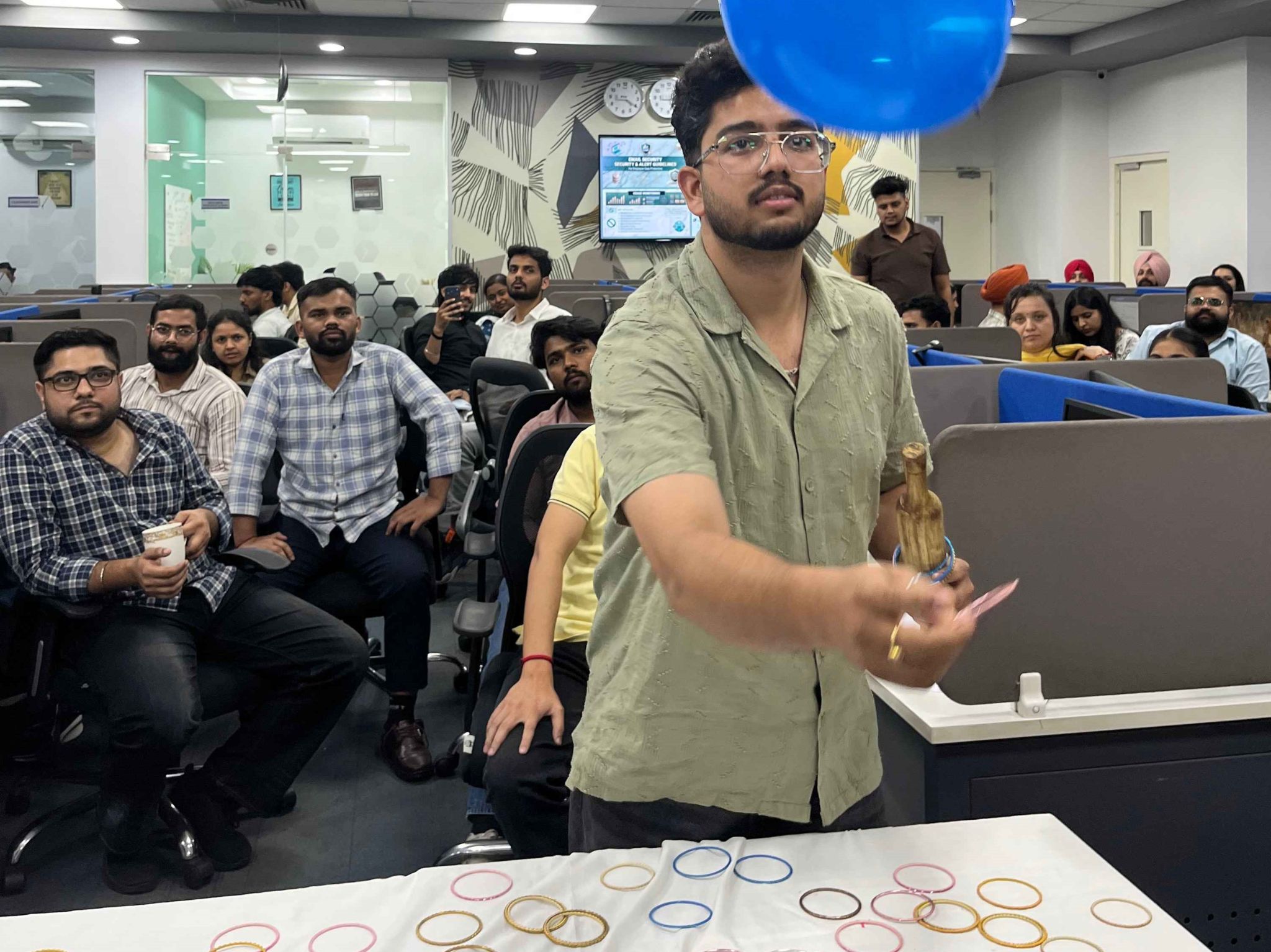 Group of coworkers seated in an office space watching a man in a light green kurta with a blue balloon overhead and a table of colorful bracelets in front.