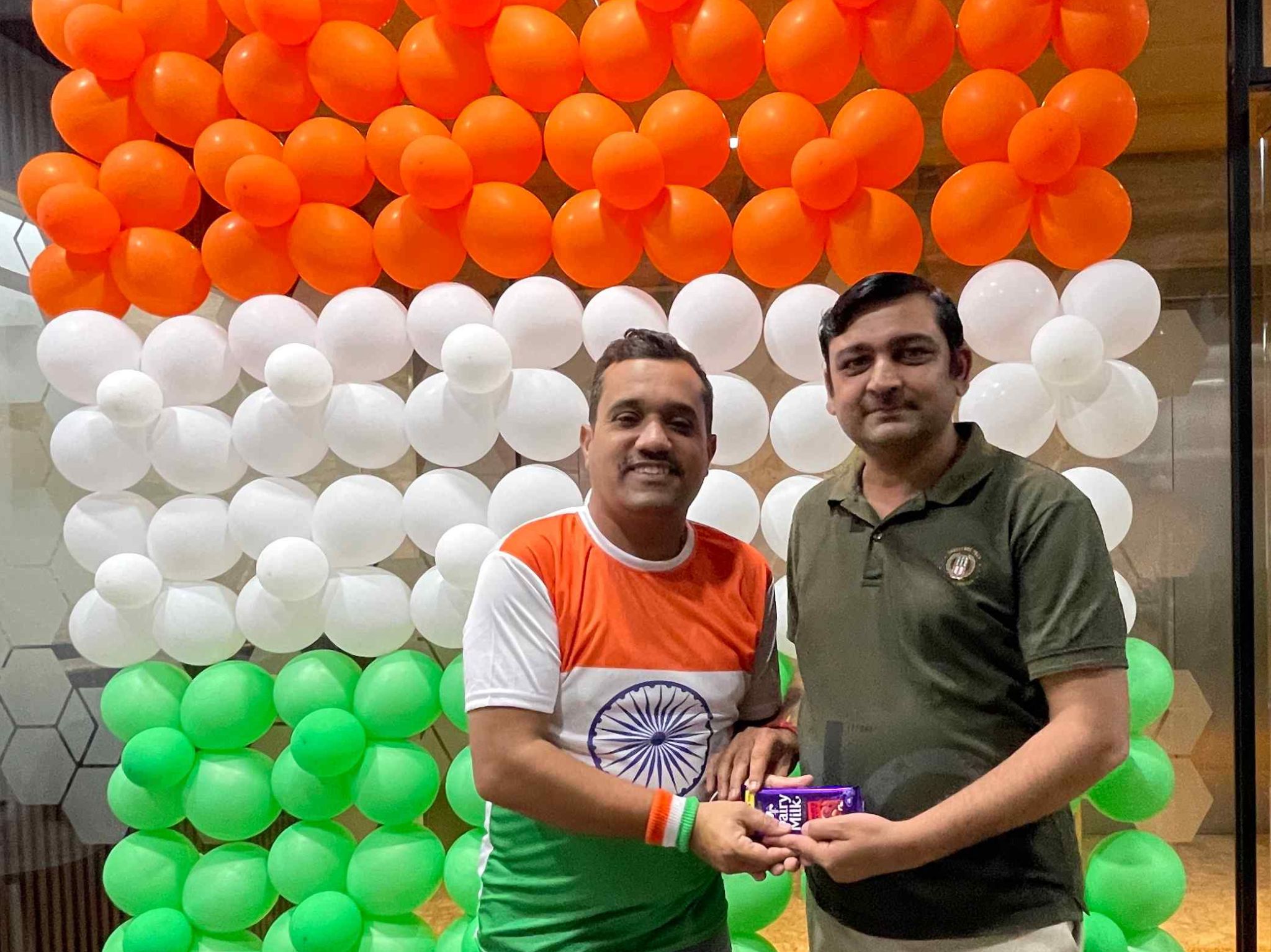 Two men posing together in front of an orange‑white‑green balloon backdrop; one wears an India jersey.